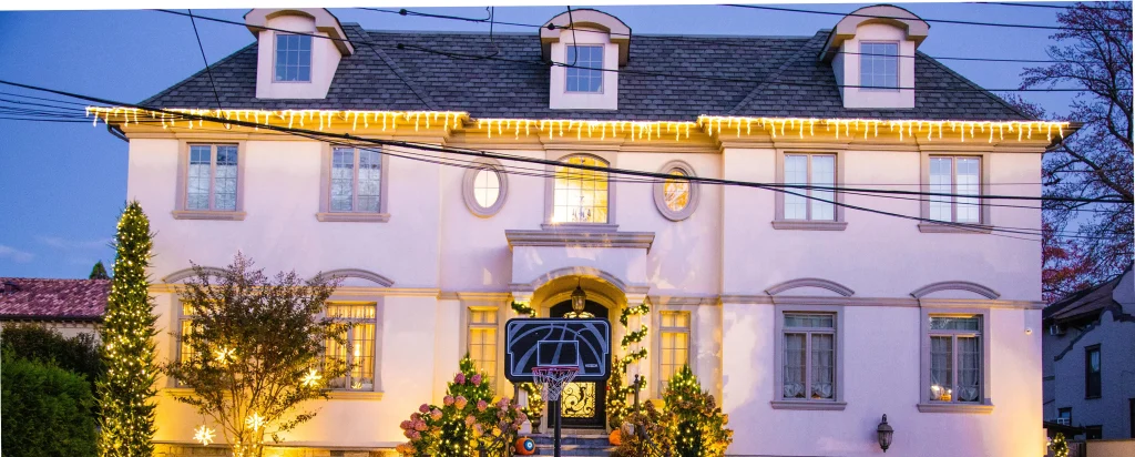 Twilight view of an elegant house brightly decorated with warm holiday lights and a basketball hoop. TITLE